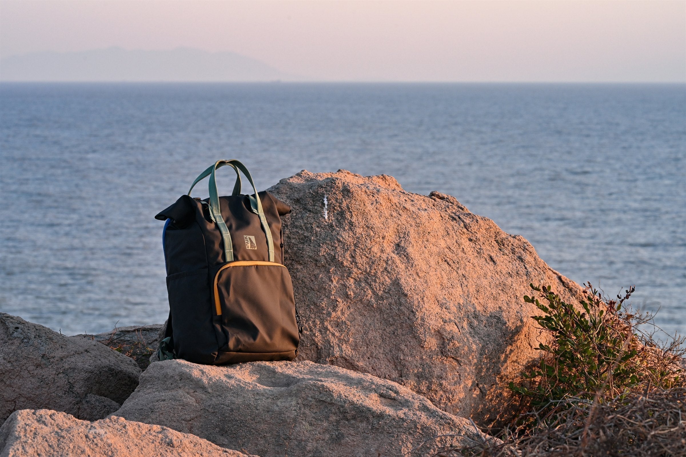 Black and olive INUK WANDER backpack standing on sunlit coastal rocks above the water